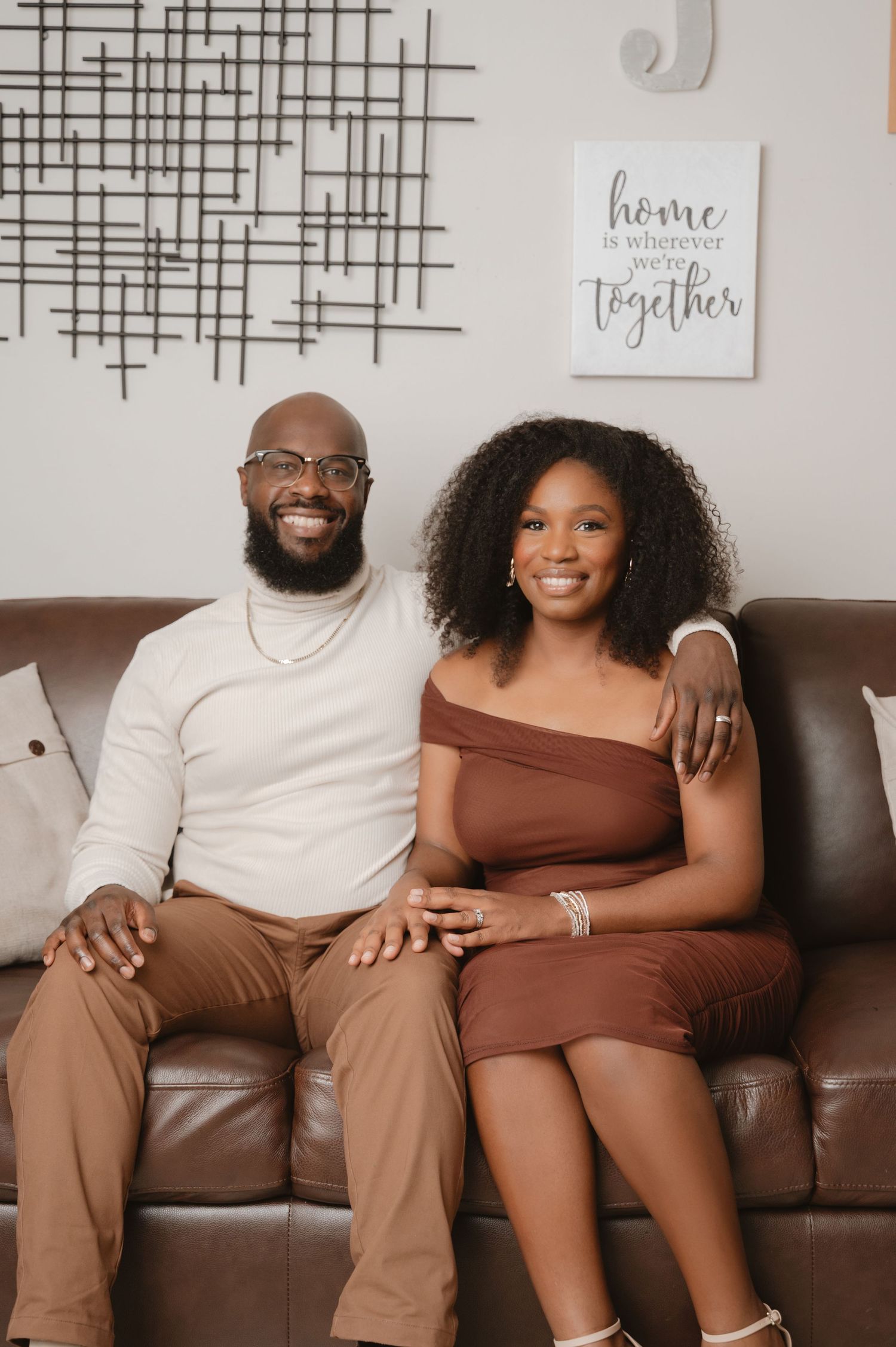 A couple sits together on a brown leather couch in a cozy living room with 'home' wall decor visible.