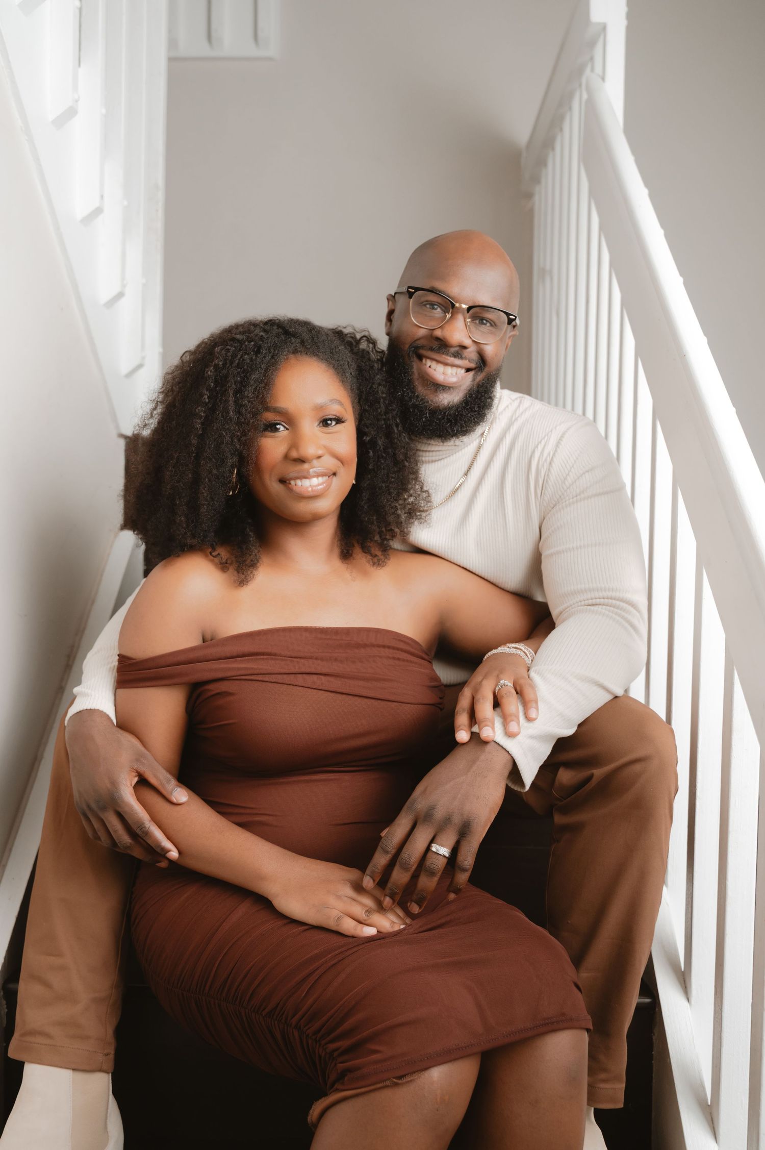 A couple in brown and white outfits pose together in a romantic maternity photoshoot against a white background.