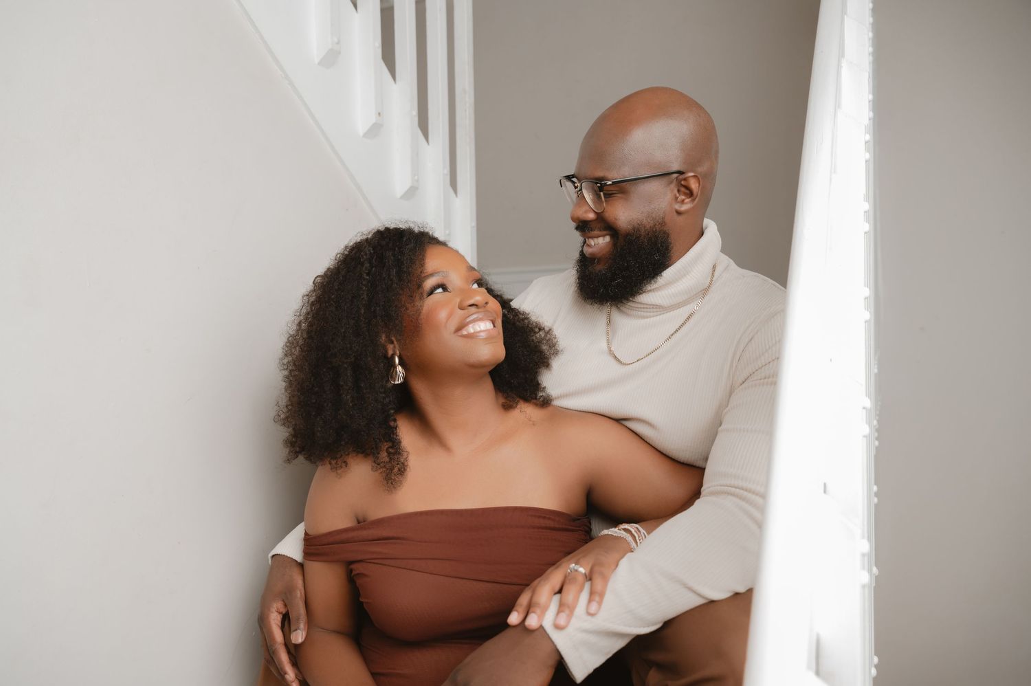 A couple shares a joyful moment together while embracing near a white wall in casual attire.