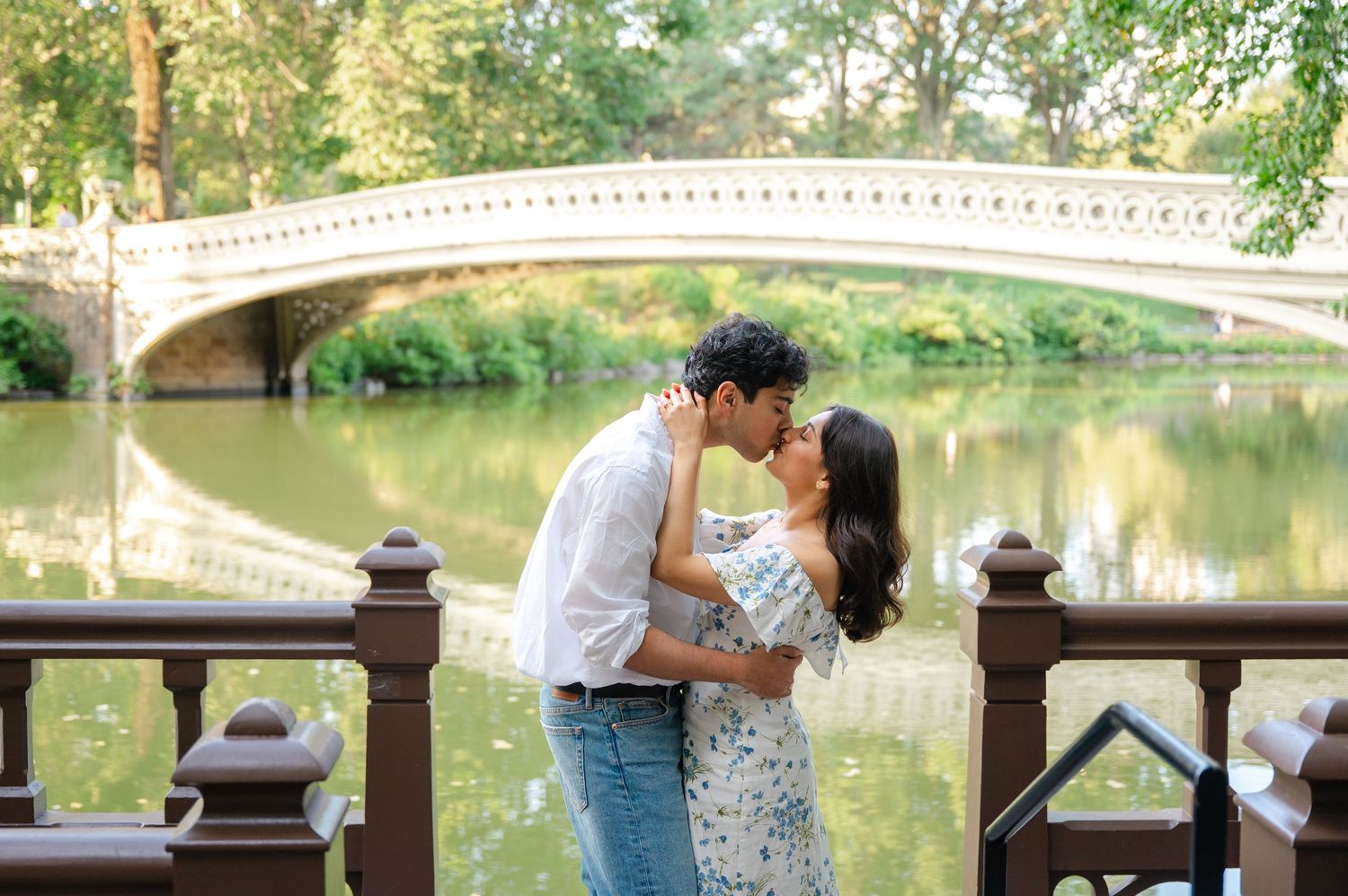 Romantic couple embraces on a scenic bridge over a tranquil pond in a lush park setting.