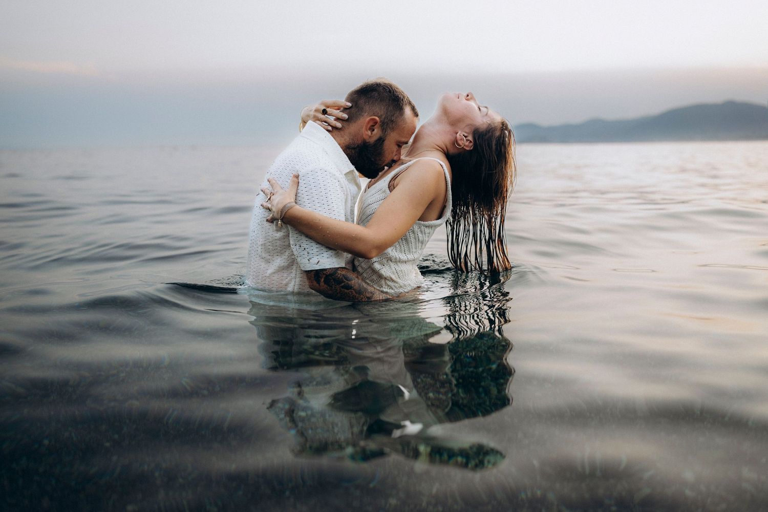 Two people embrace while floating in calm ocean waters at sunset with mountains visible in the distance.