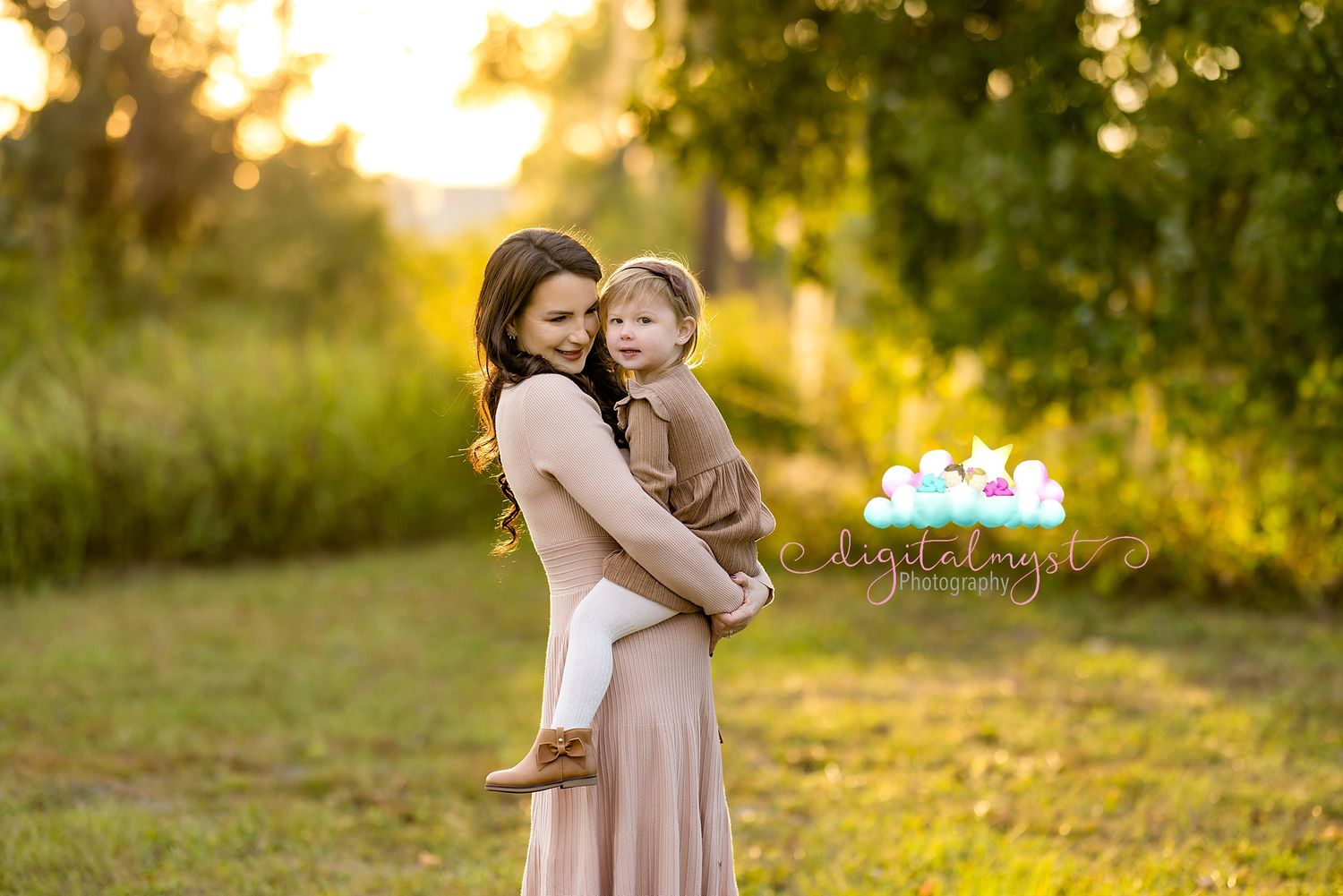 Two people embrace in a golden sunlit field during sunset while wearing matching mauve dresses.