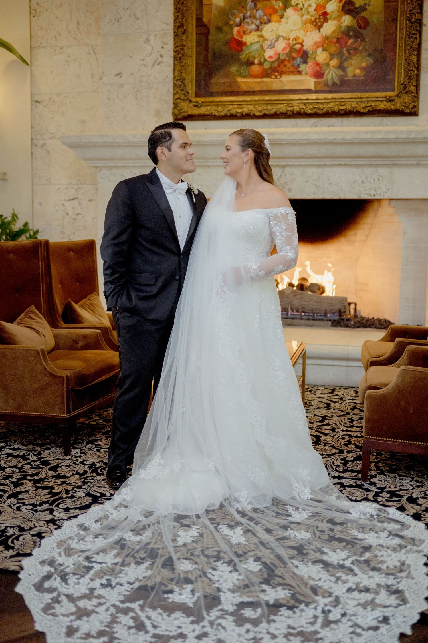 A bride in a flowing white gown with an elaborate train poses with her groom in an elegant indoor setting.