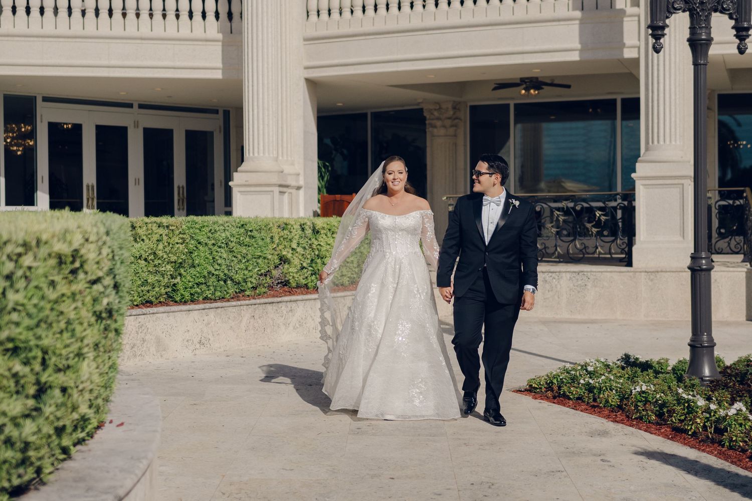 A bride and groom walk together on a paved path surrounded by manicured gardens and elegant architecture.