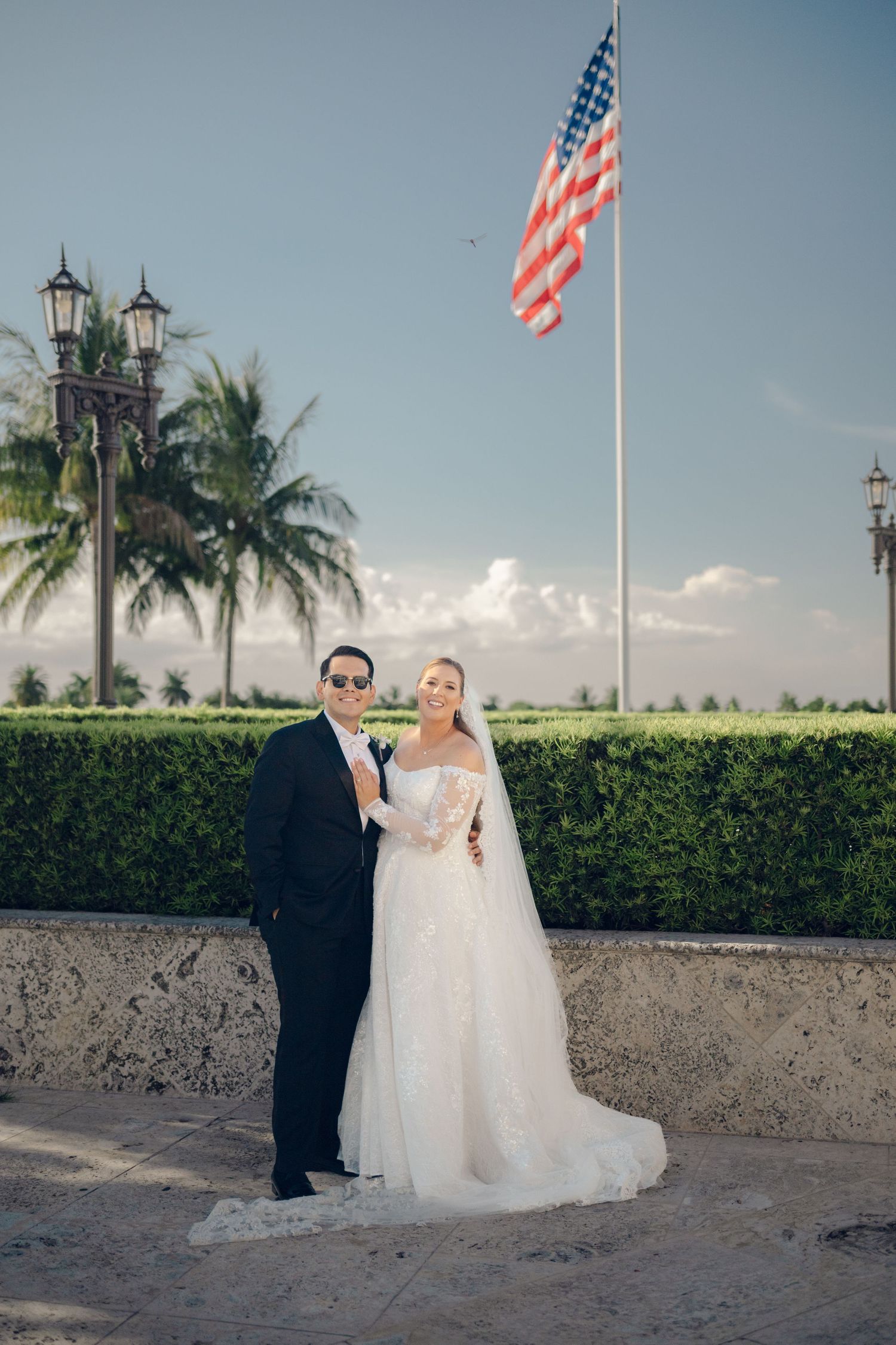 Wedding couple poses against stone wall with American flag and palm trees under blue sky.