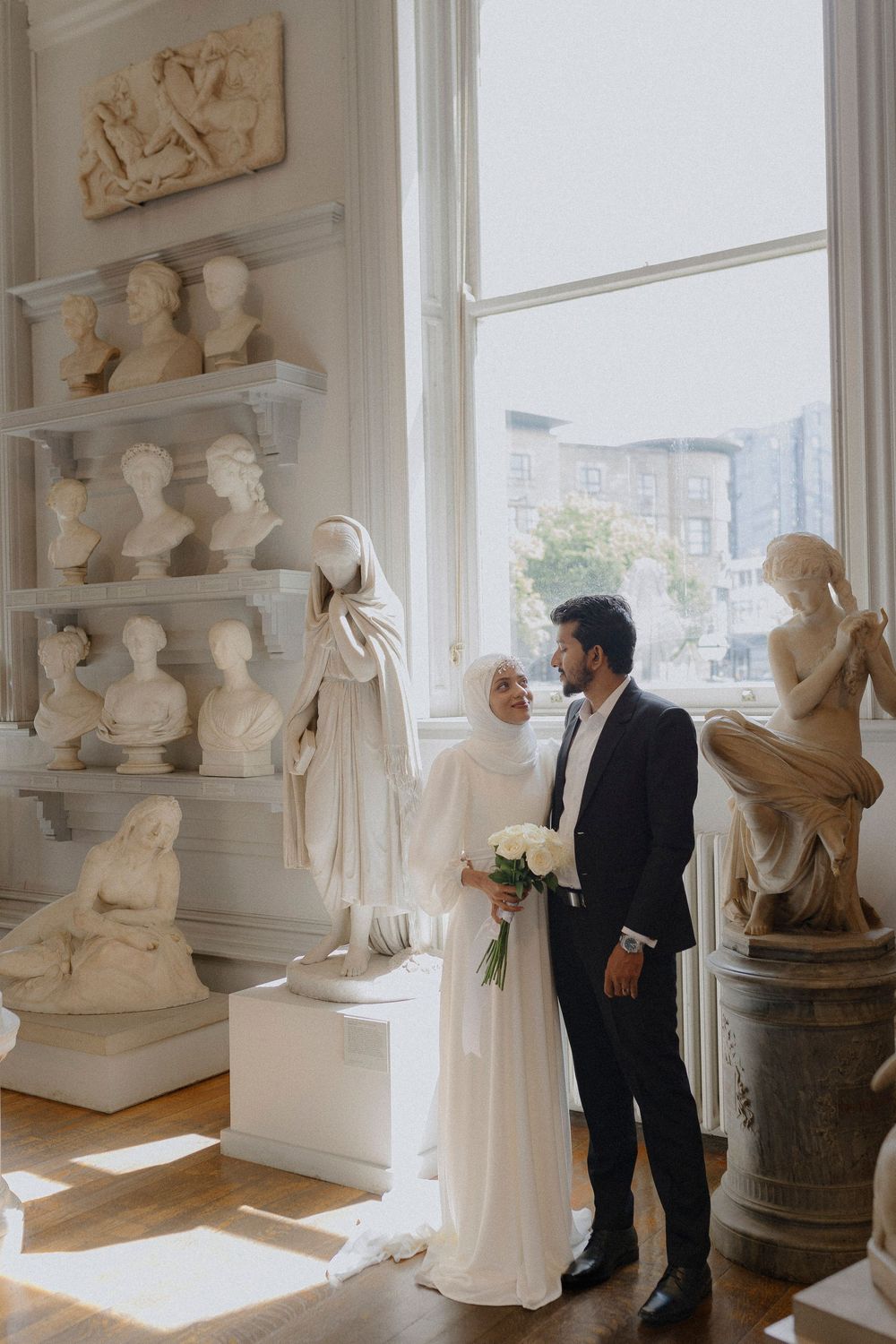 A couple poses in an elegant room filled with classical busts and sculptures on display shelves near bright windows.