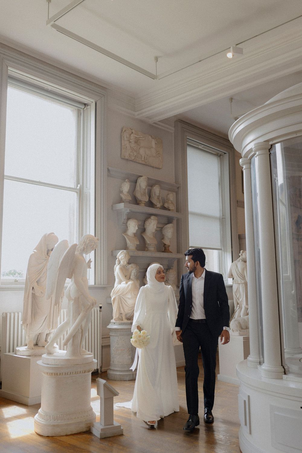 Art gallery interior with white marble sculptures and statues displayed along bright windowed hallway.
