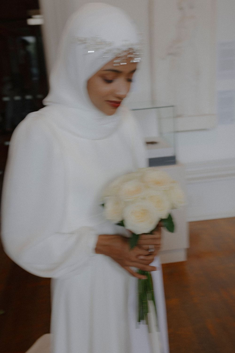 A bride in a flowing white dress and hijab holds a bouquet of cream-colored roses.