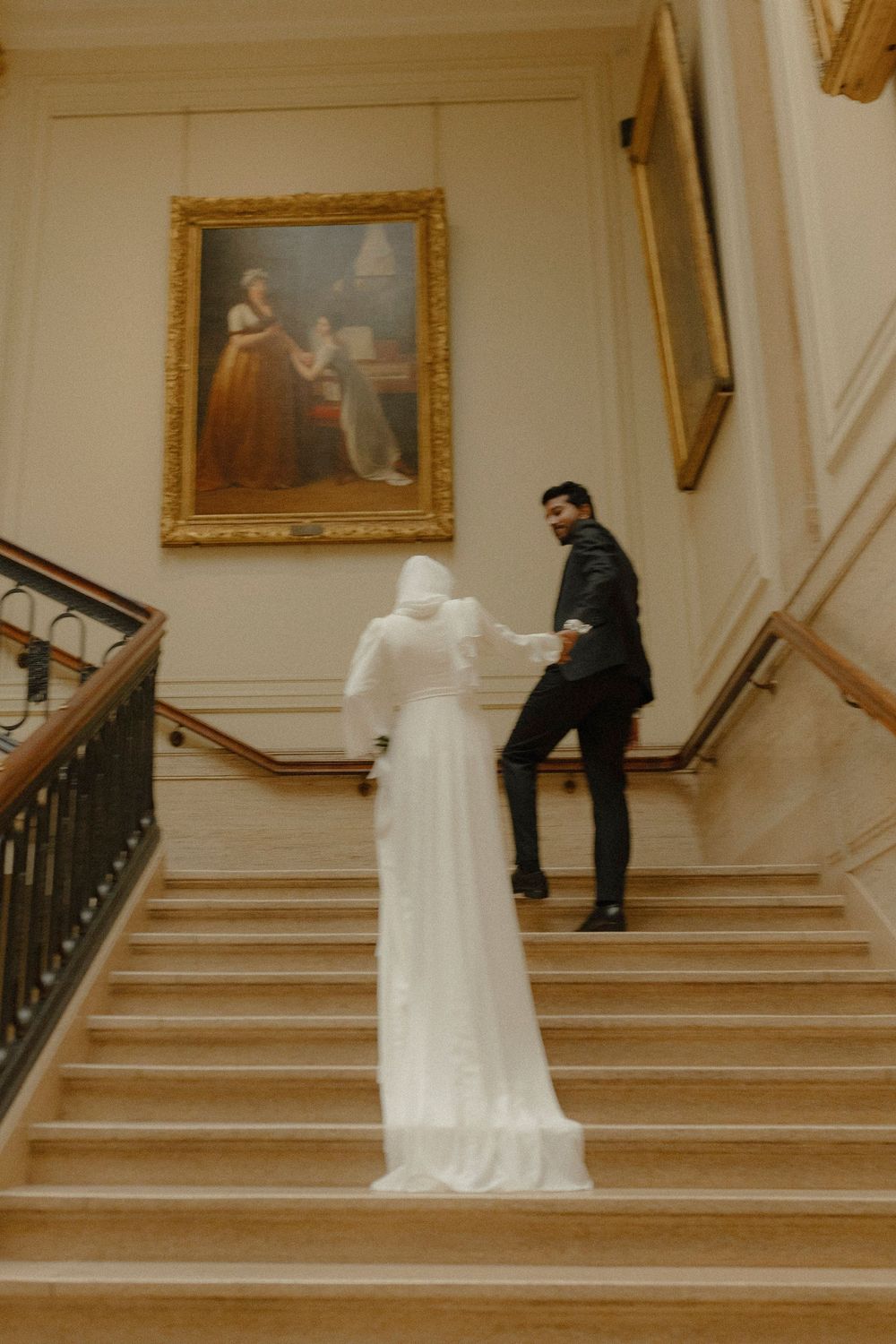 A couple descends an elegant staircase with gold-framed artwork on the walls.