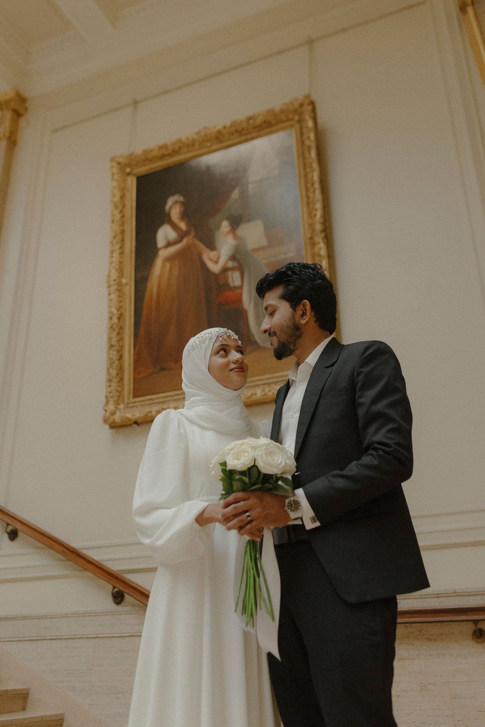 A couple in a white dress and dark suit embrace in an elegant room with ornate gold-framed paintings on the wall.
