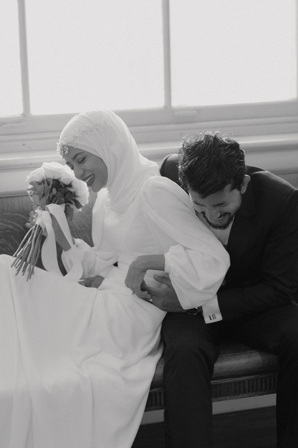 A black and white photo of a couple sharing a joyful moment on their wedding day, both laughing together.