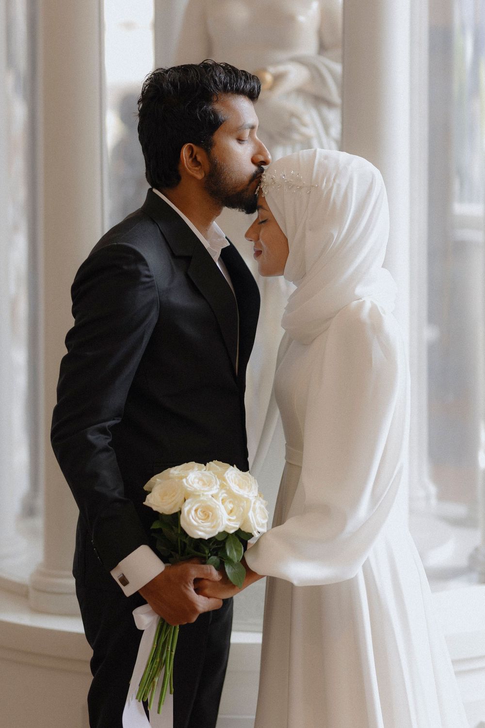 A couple in formal wedding attire share a tender moment, with the groom in black and bride in white holding roses.