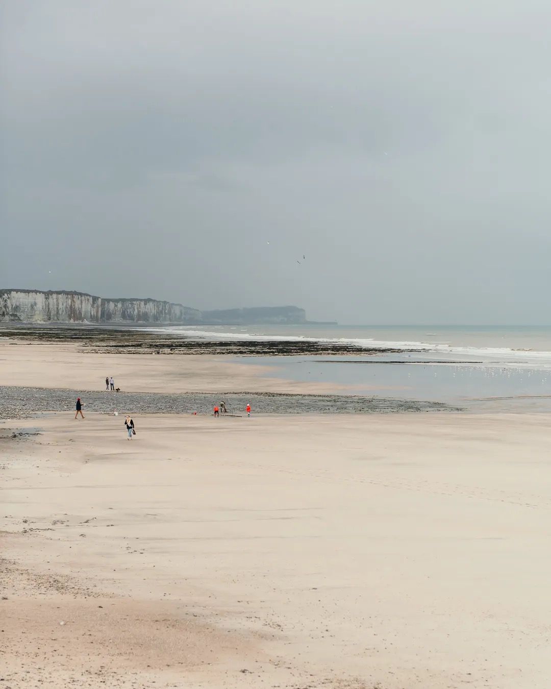 Wide sandy beach on a cloudy day with distant cliffs and gentle waves.