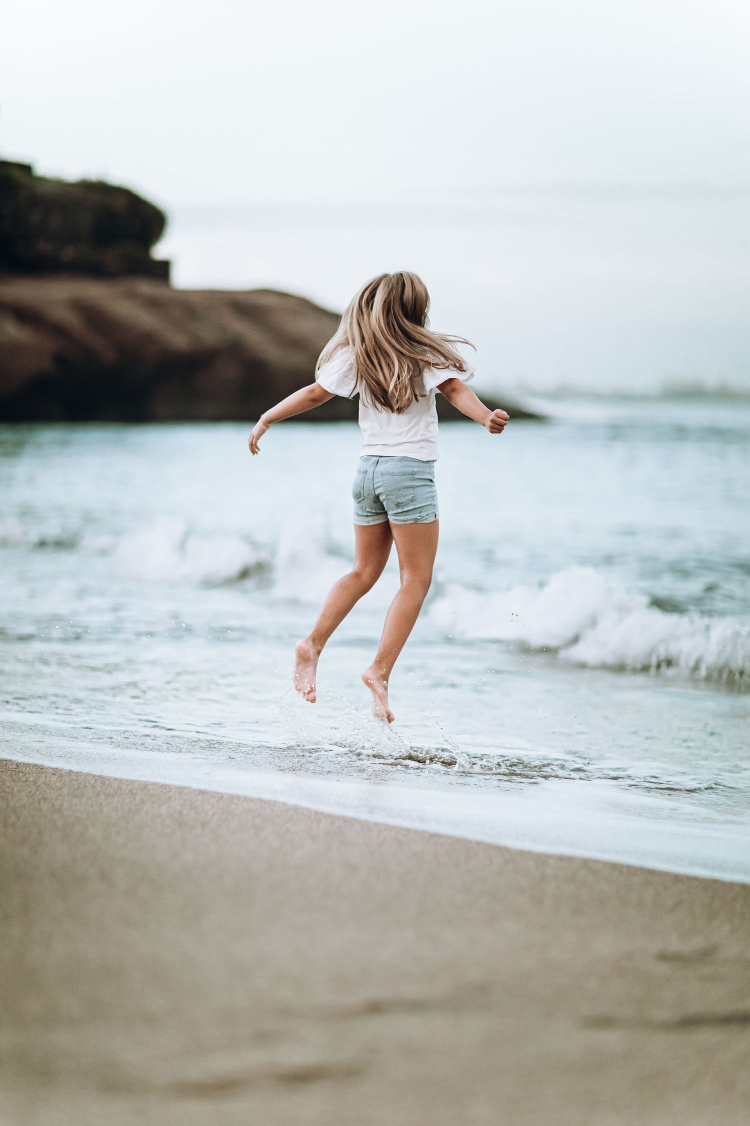 Person jumping joyfully on a beach with waves and rocks in the background.
