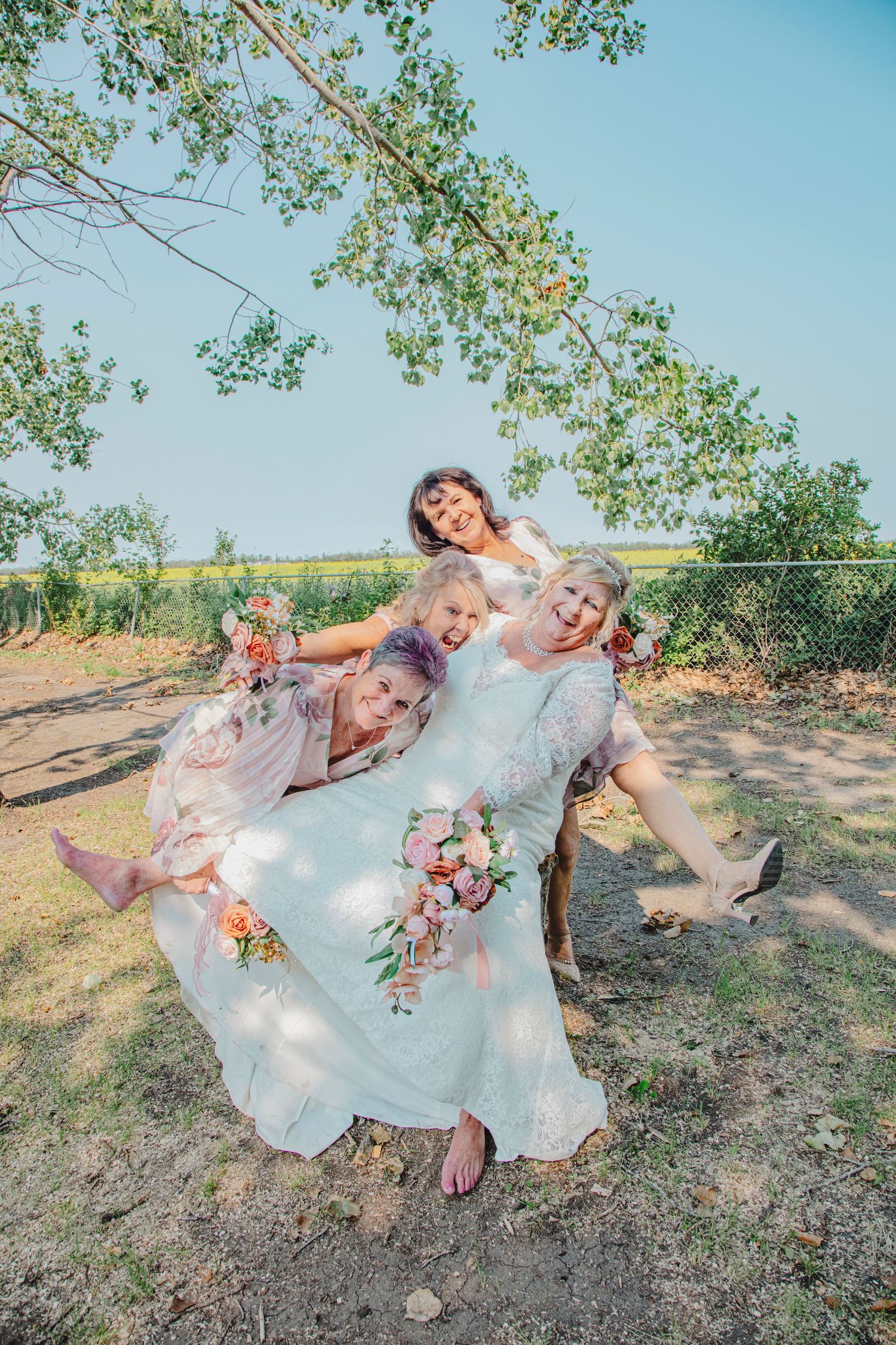 Bride in white gown laughing with bridesmaids in blush dresses holding floral bouquets outdoors on a sunny day.