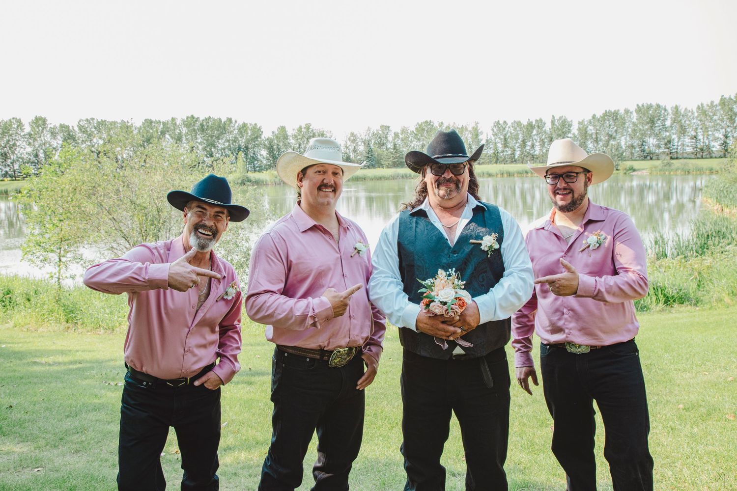 Four groomsmen in pink shirts and black cowboy hats pose outdoors near a pond at a country western wedding.