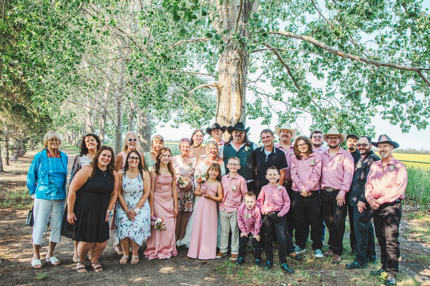 Large family group photo outdoors under trees at a country wedding with guests in pink and western attire.