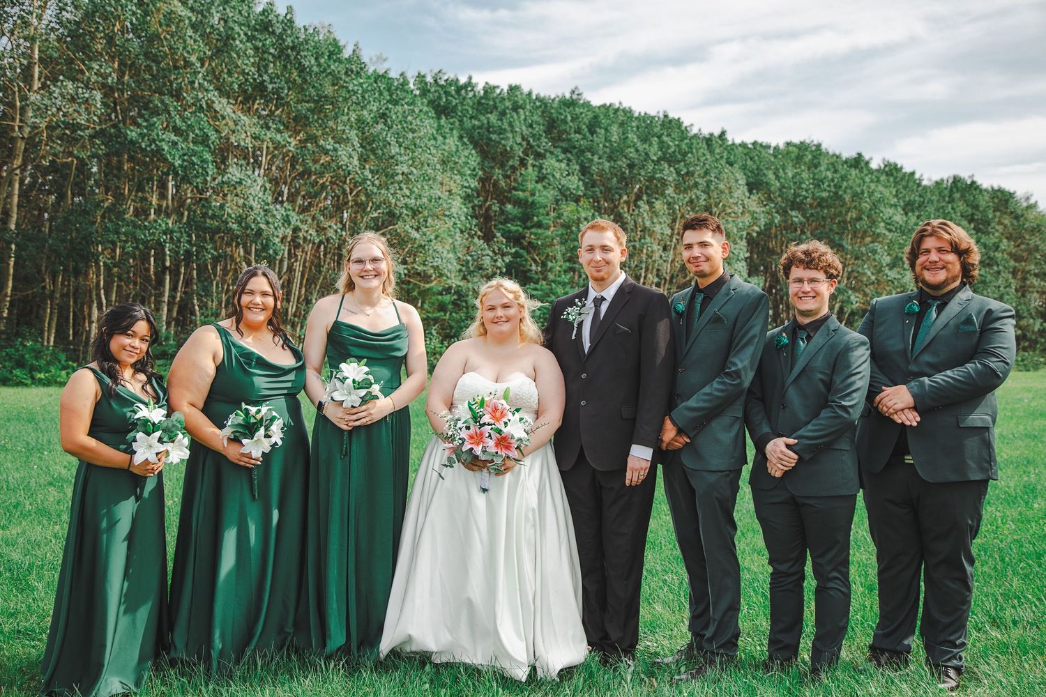 Wedding party in emerald green attire poses outdoors with bride and groom on lush green lawn with a pine forest in background.