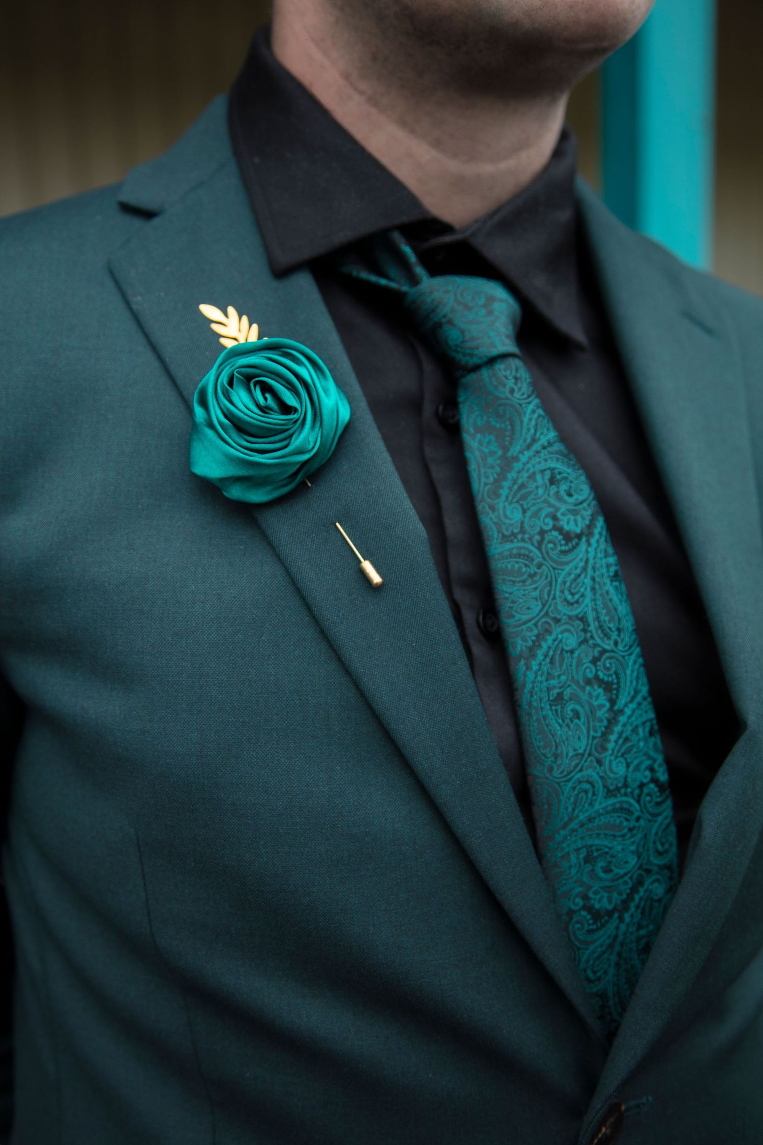 Man wearing green suit with matching paisley tie and teal rose boutonniere with gold leaf pin.
