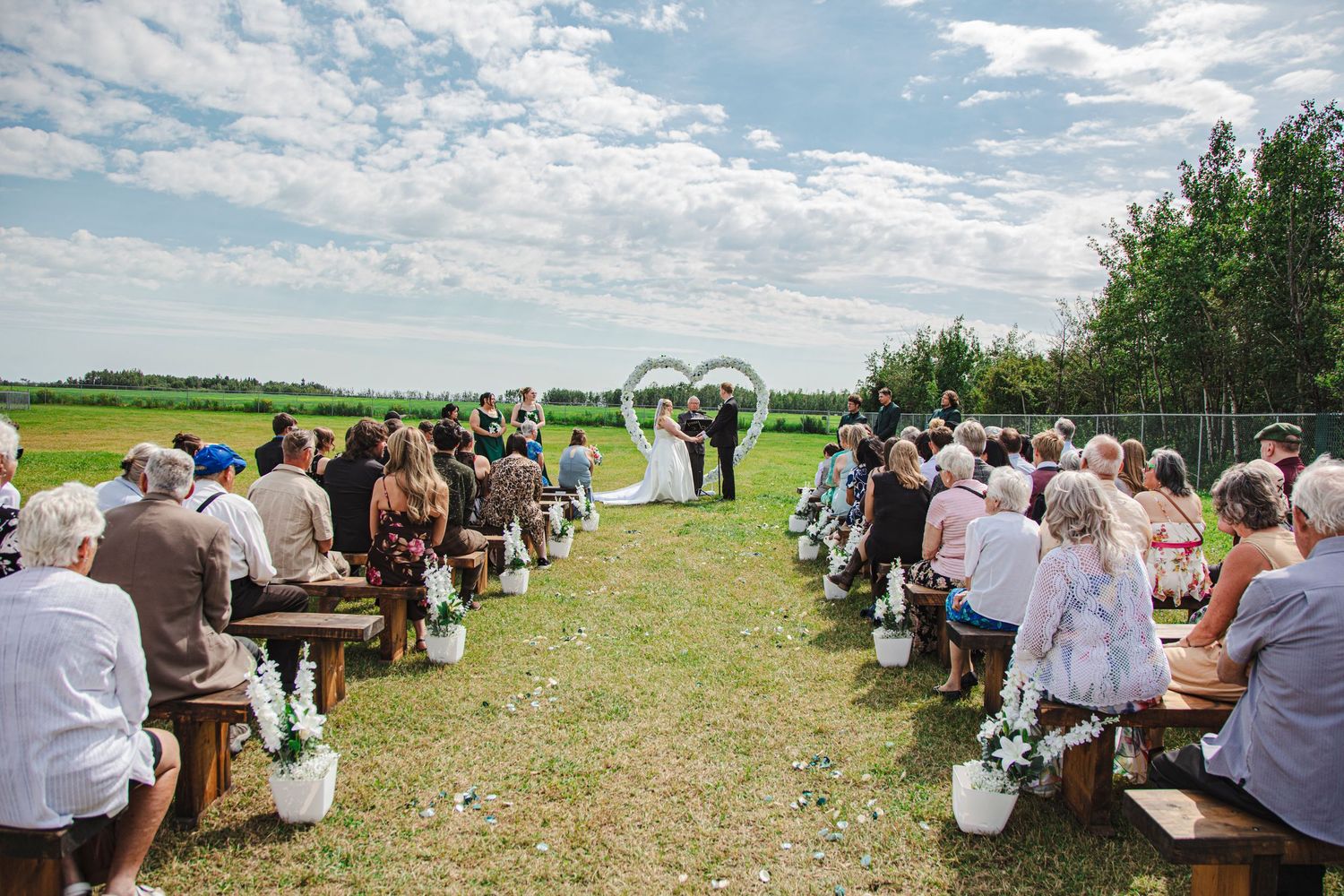 Outdoor wedding ceremony on green lawn with guests seated on wooden benches flanking a floral arch aisle.