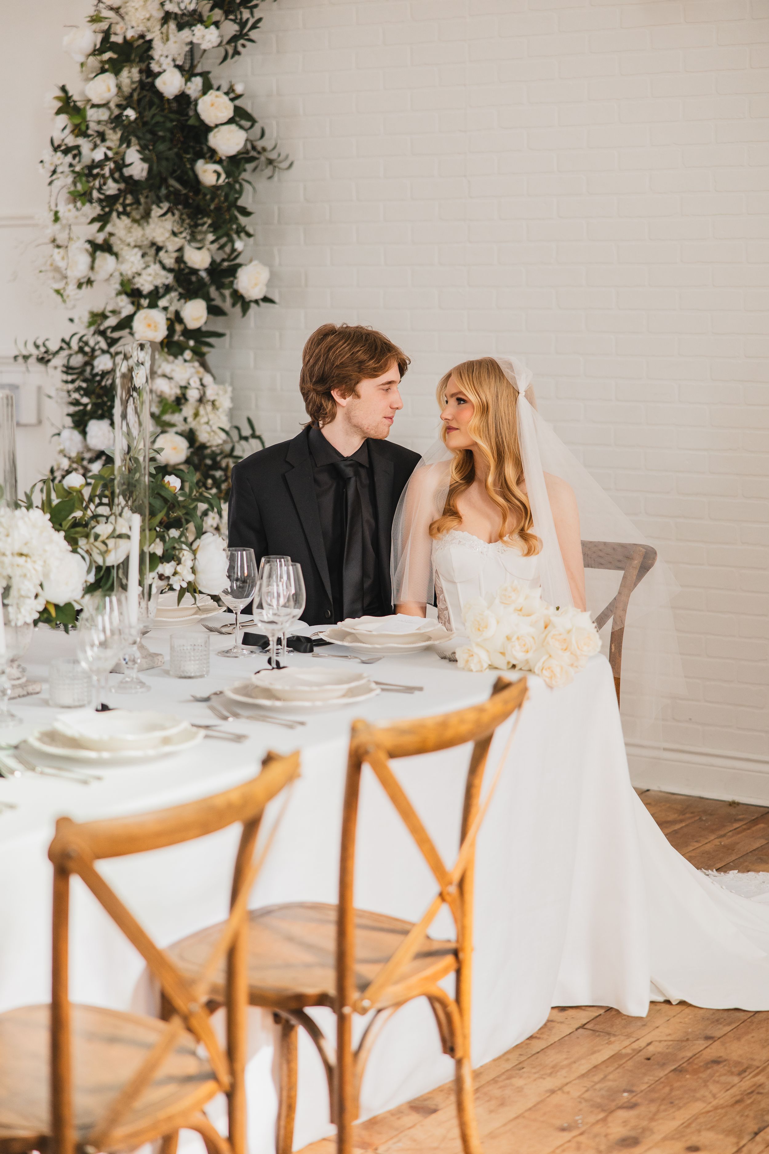 A bride and groom sit at wedding reception table with white linens, wooden chairs and floral arrangements in a bright indoor space.