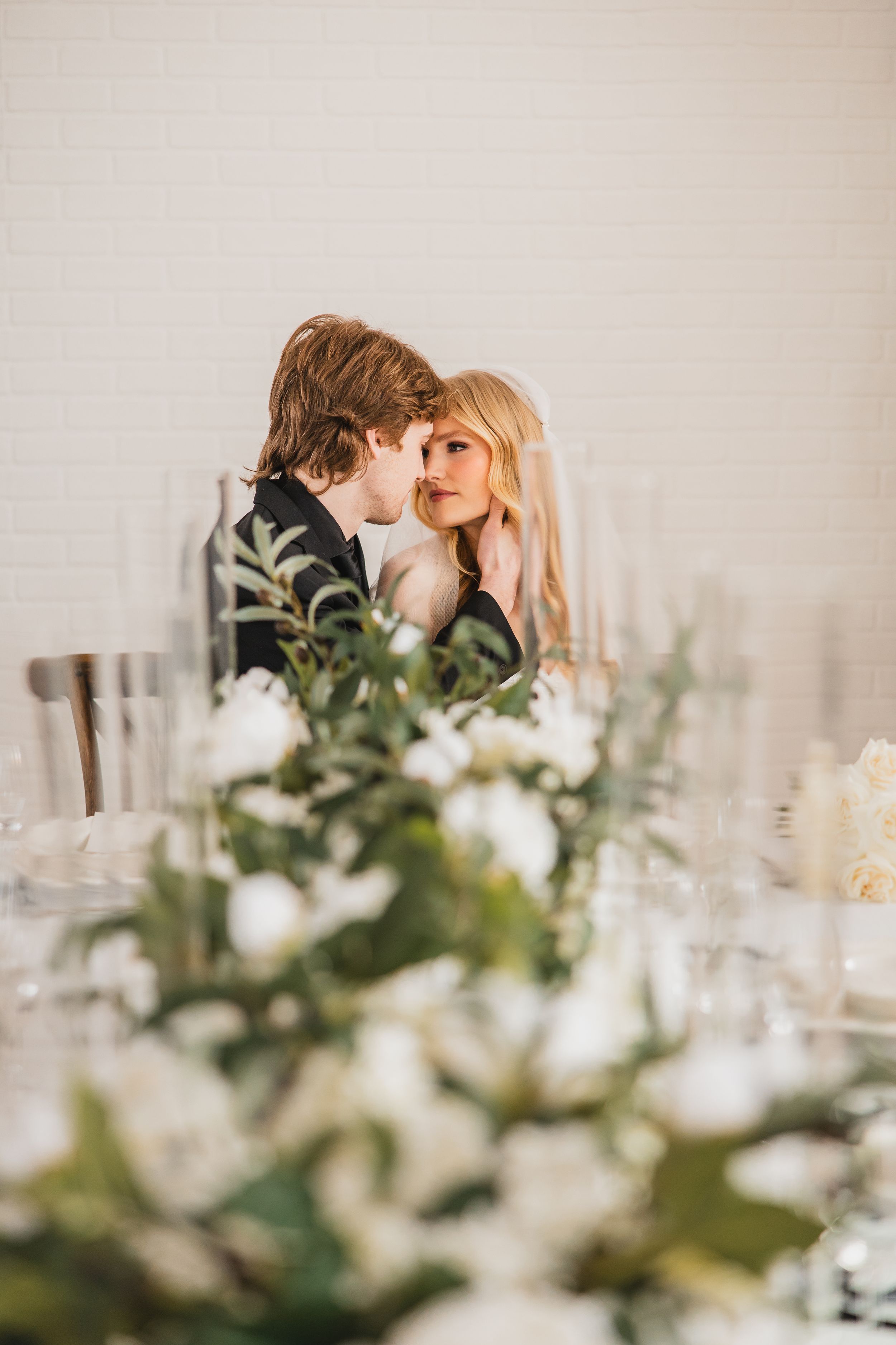 A romantic moment captured at an elegant wedding reception with white floral centerpieces and greenery in the foreground.