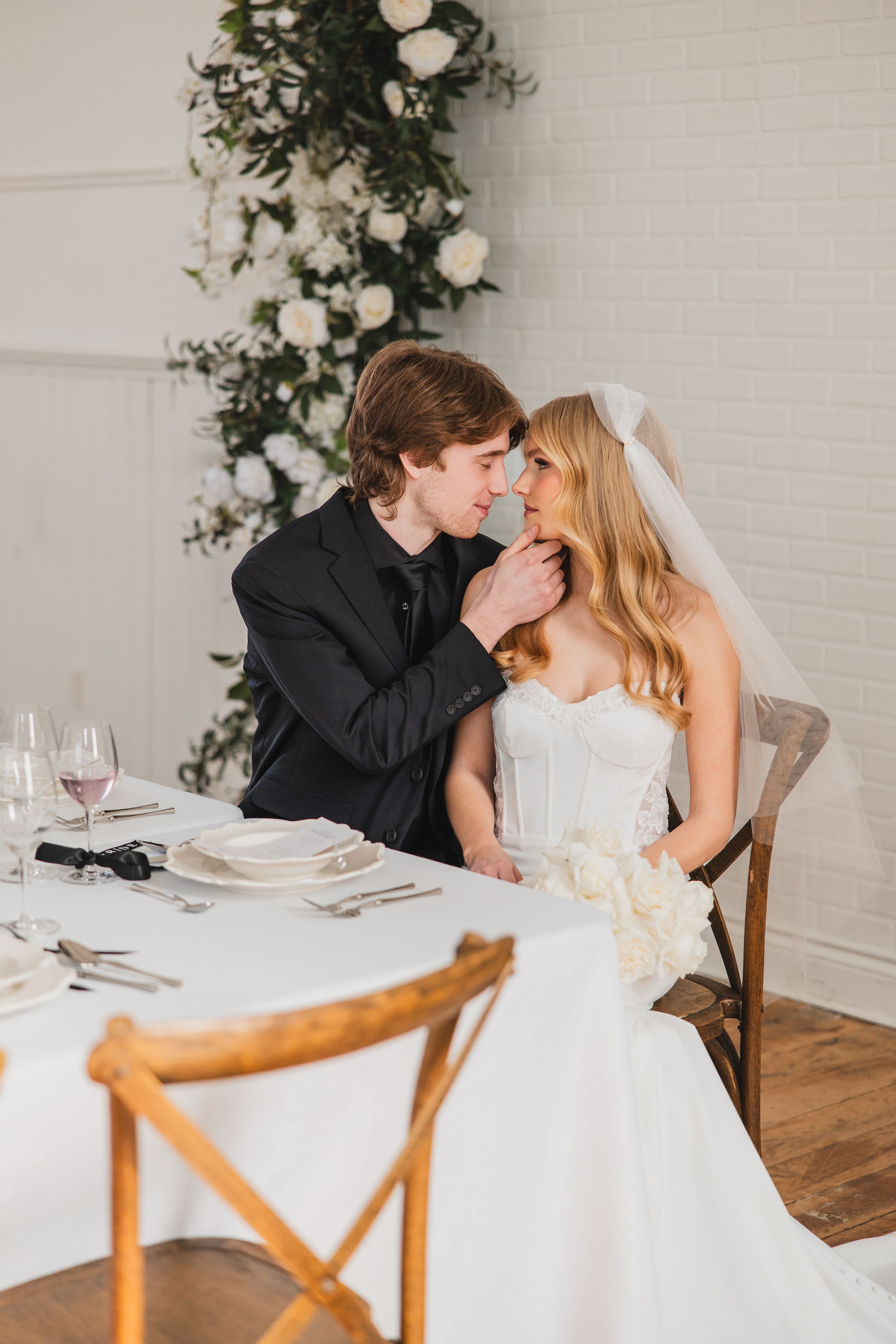 A romantic wedding couple shares intimate moments at a white-clothed dining table with elegant place settings and floral decor.