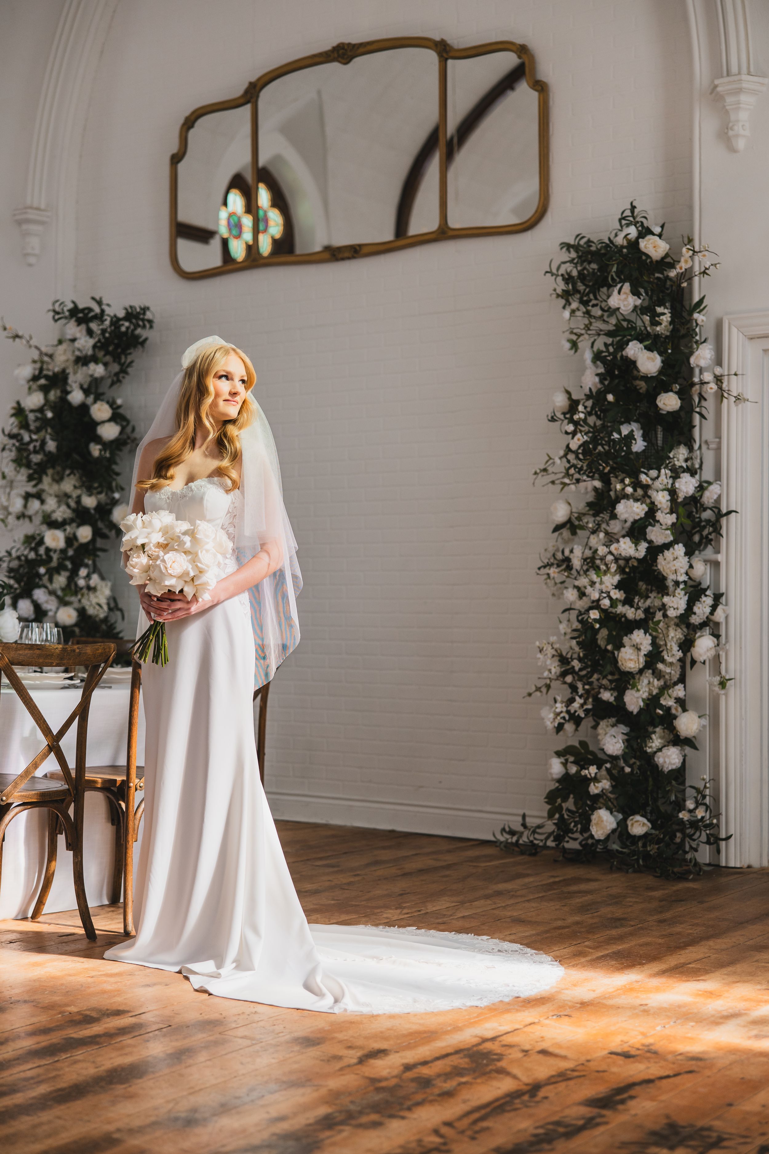 An elegant bridal portrait with white floral arrangements, a vintage mirror, and a flowing wedding gown in a bright indoor setting.