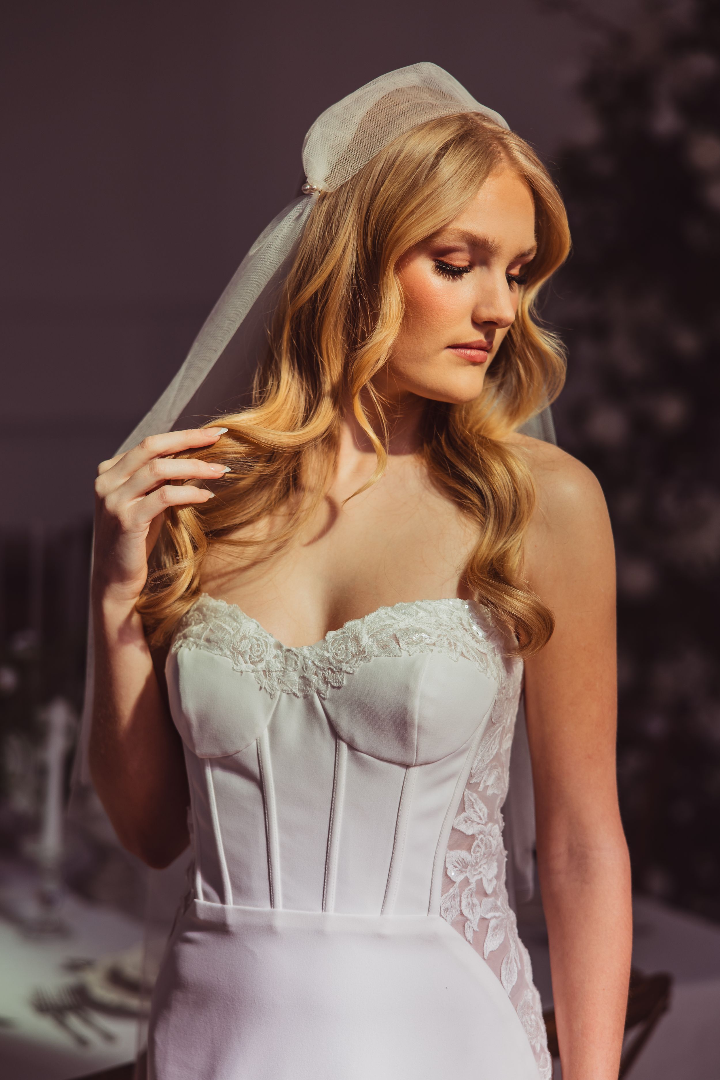 A bride in a white corset wedding dress with lace details and flowing veil poses elegantly against a dark background.