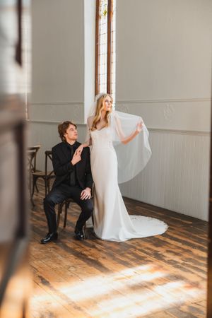 A couple in formal wedding attire poses together in a sunlit chapel with hardwood floors and white walls.