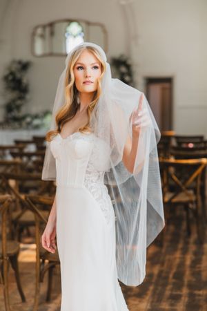 A bride in a flowing white wedding gown and veil poses in a rustic indoor venue with wooden floors and chairs.