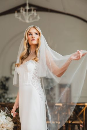 Elegant bride in form-fitting lace gown with flowing veil poses gracefully in front of arched window and chandelier.