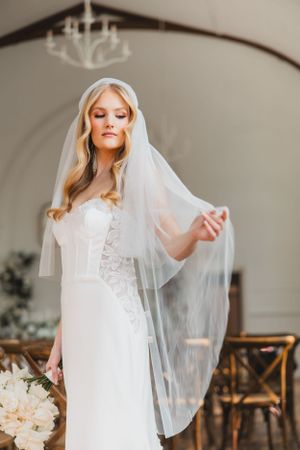 A bride in an elegant white lace wedding dress and flowing veil poses gracefully in a light-filled room.
