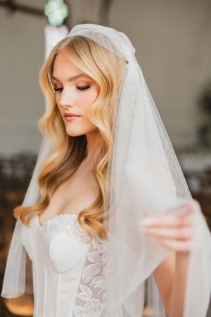 A bride wearing a delicate white veil and lace corset wedding dress with soft wavy blonde hair and romantic makeup.