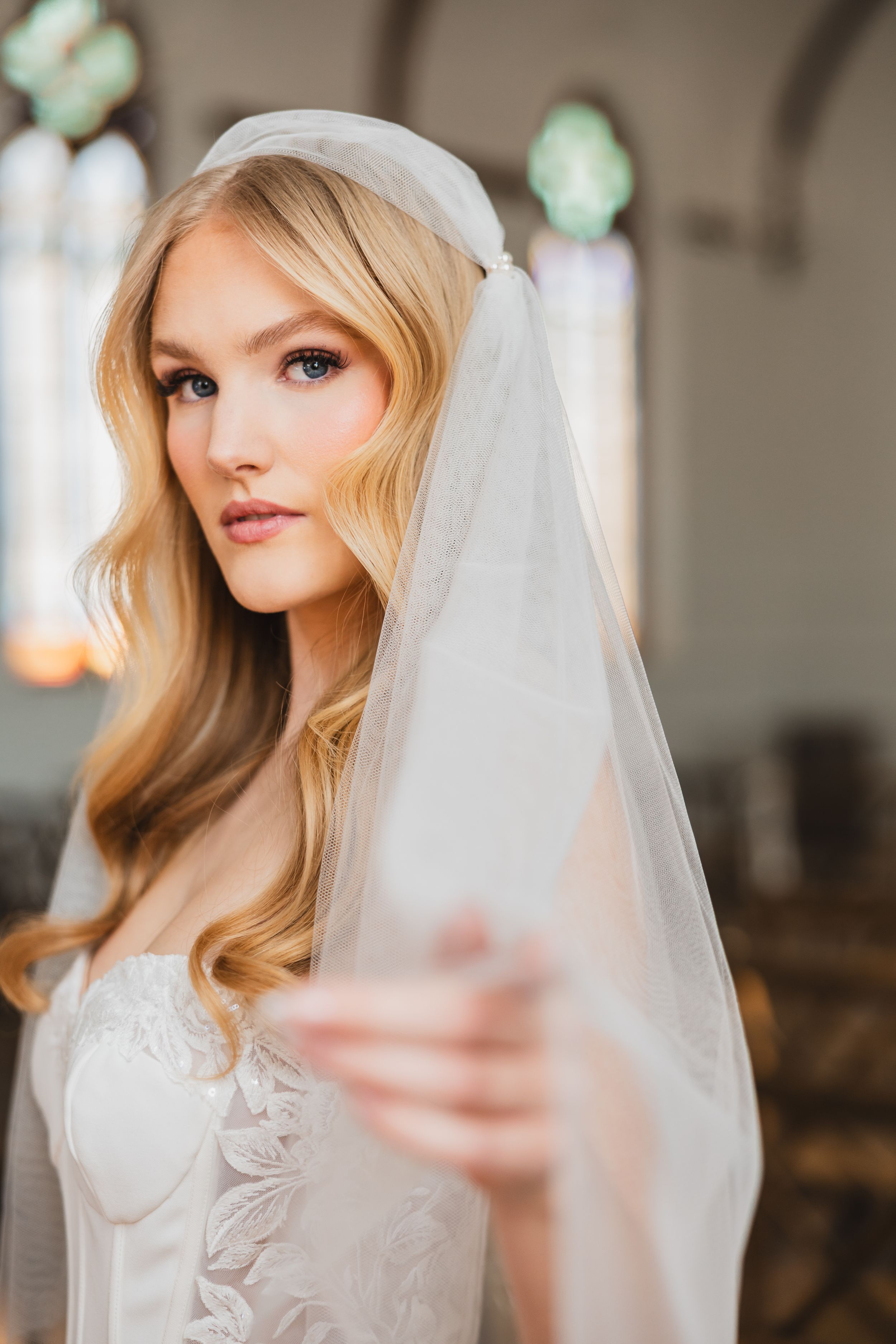 Bride wearing white lace wedding dress and sheer veil with soft wavy blonde hair styled elegantly.