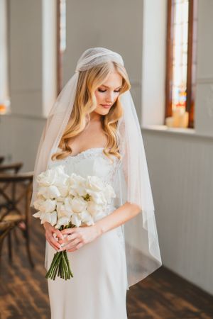 Elegant bride in white strapless gown holds classic white tulip bouquet while standing in sunlit church.