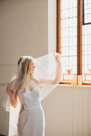 Bride in white dress stands by sunlit church windows with flowing veil creating ethereal atmosphere.