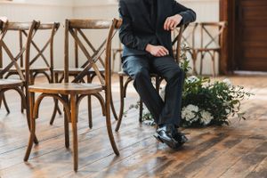 A groom waiting for his bride on a wooden crossback chair at the wedding venue with hardwood floors against a neutral wall.