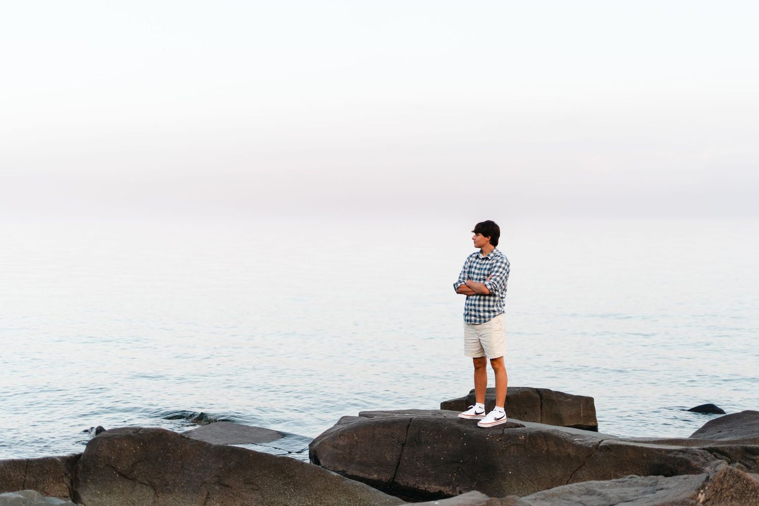 Person standing on coastal rocks overlooking a misty ocean at sunset while wearing casual summer clothing.