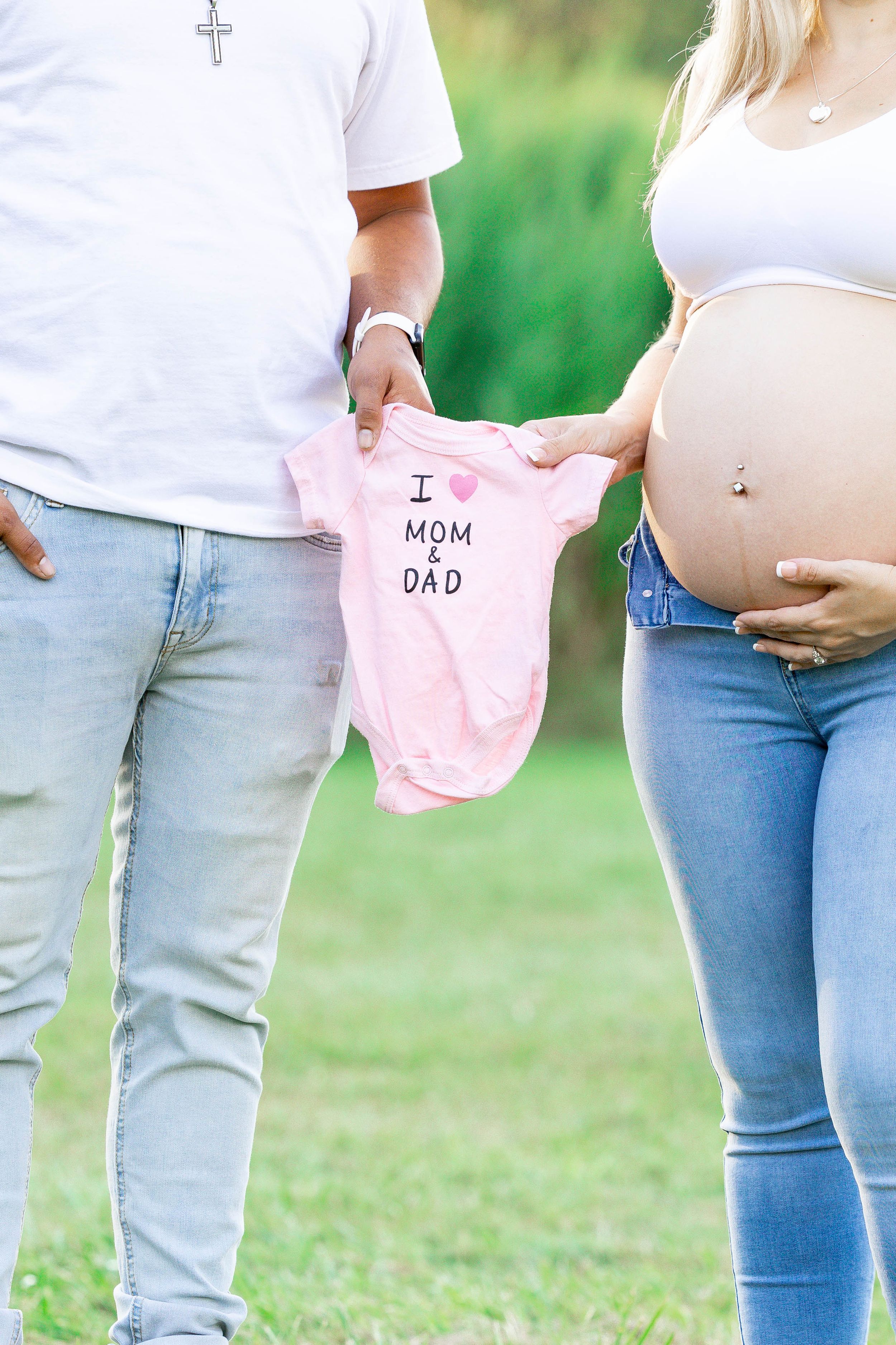 An expectant couple holds a pink baby onesie with 'I love mom & dad' text in a sunlit outdoor setting.
