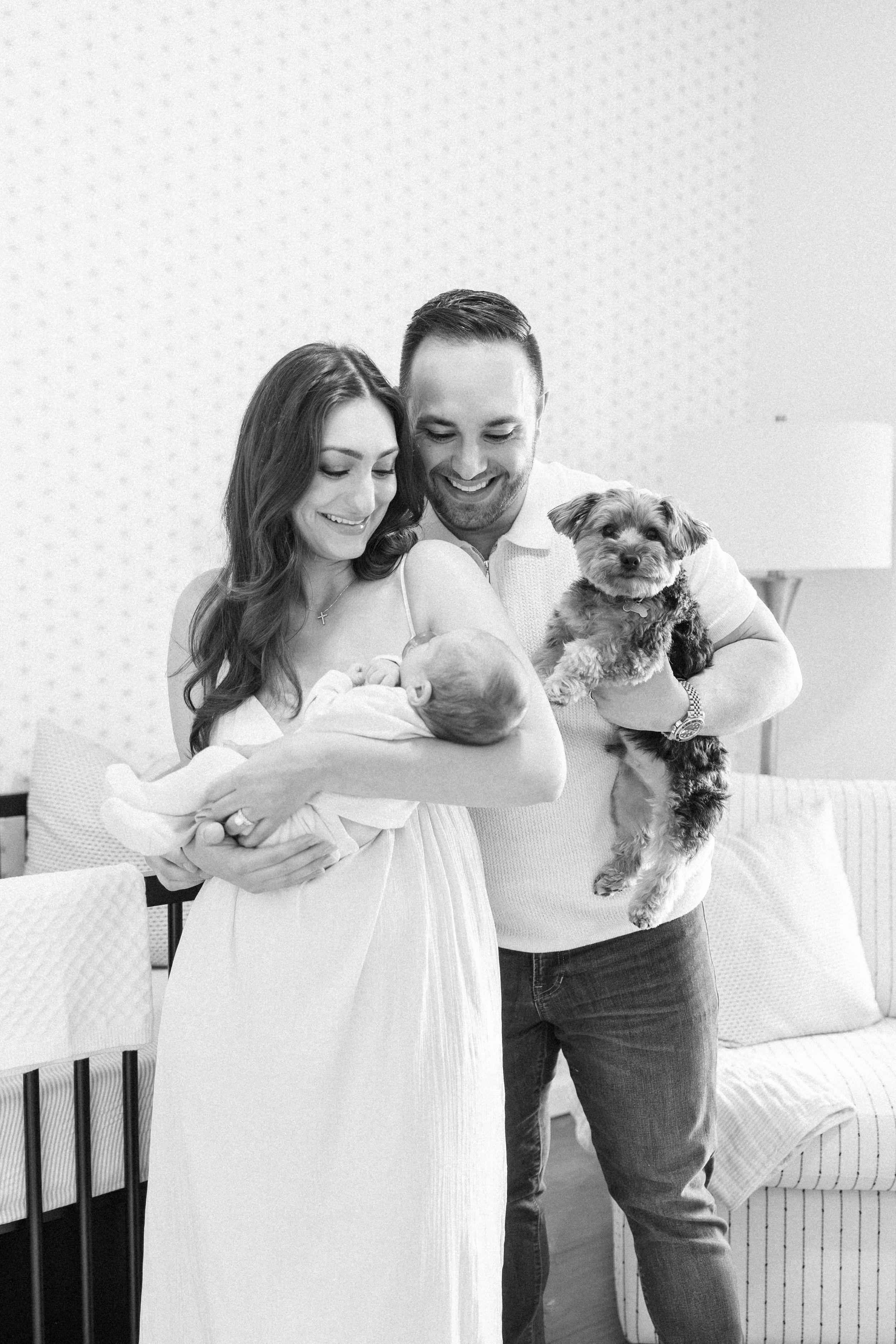 Black and white photo of couple holding their newborn baby and small dog in a bright white bedroom.