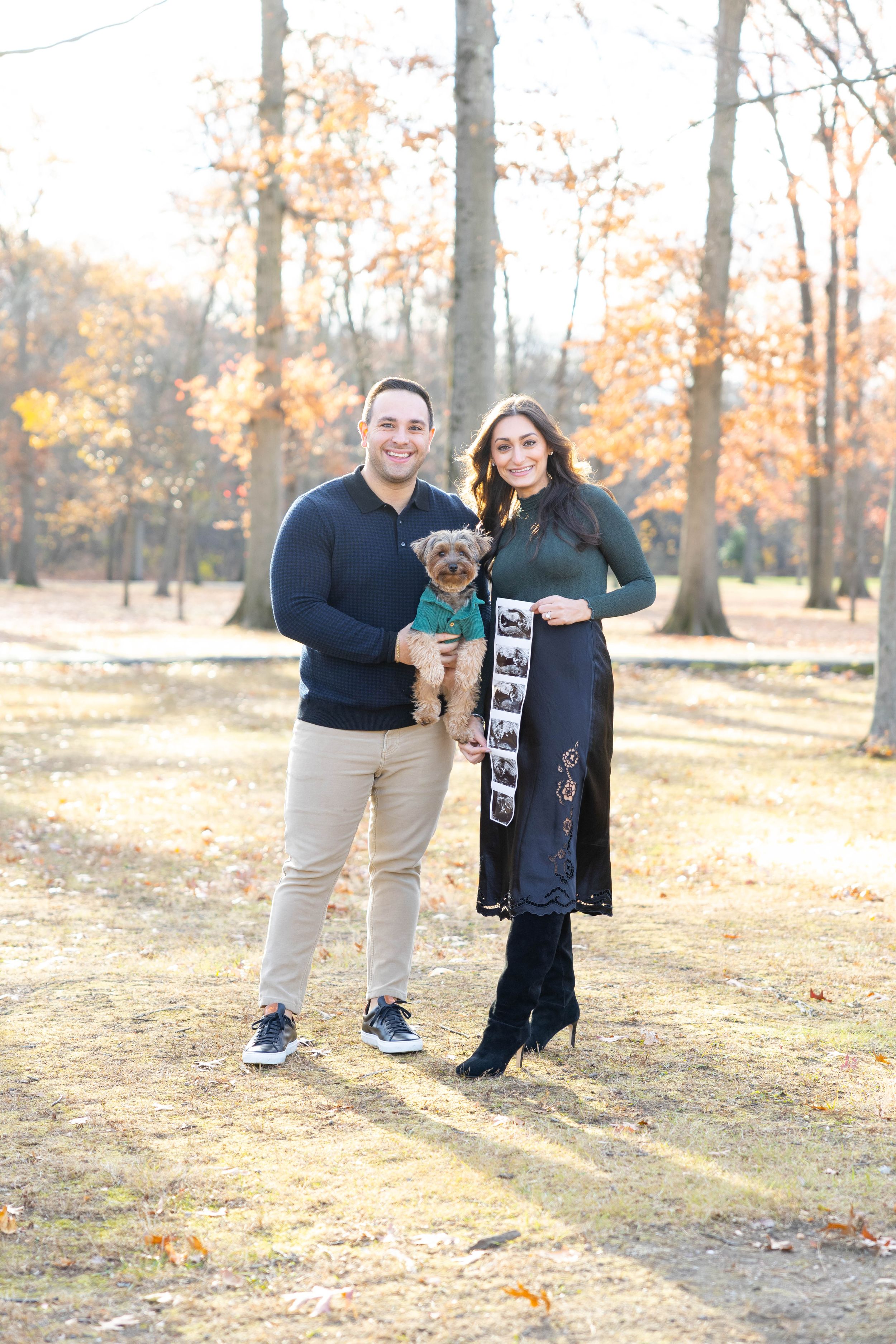 A couple and their small dog pose together in a park during fall with golden leaves in the background.