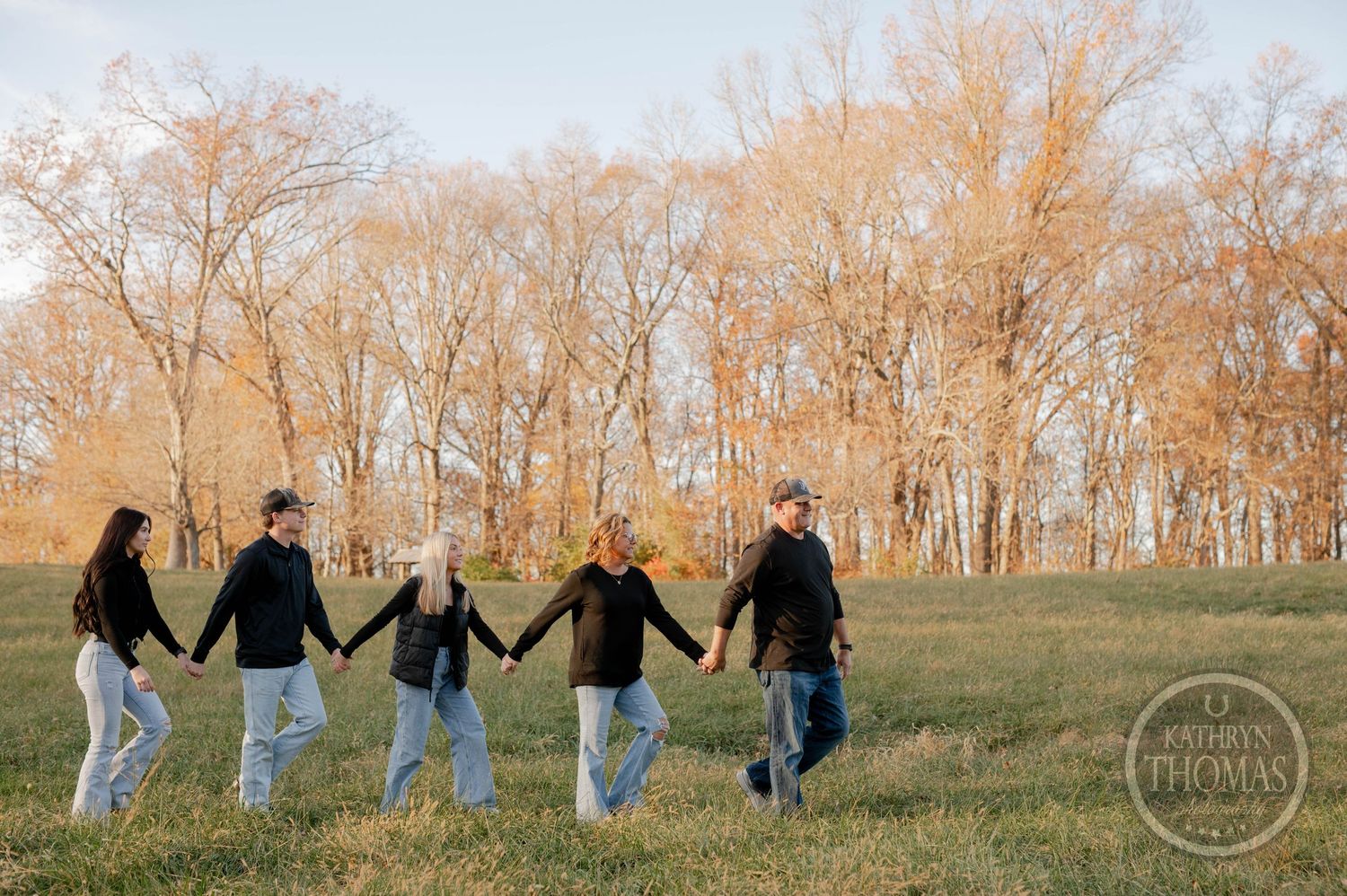 A group of people holding hands while walking through an autumn field with trees in the background.