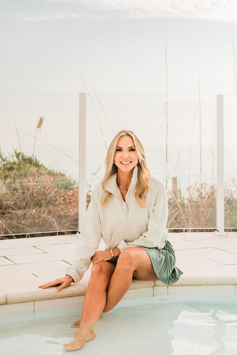A person in a cream sweater and patterned skirt sits poolside on a bright sunny day.