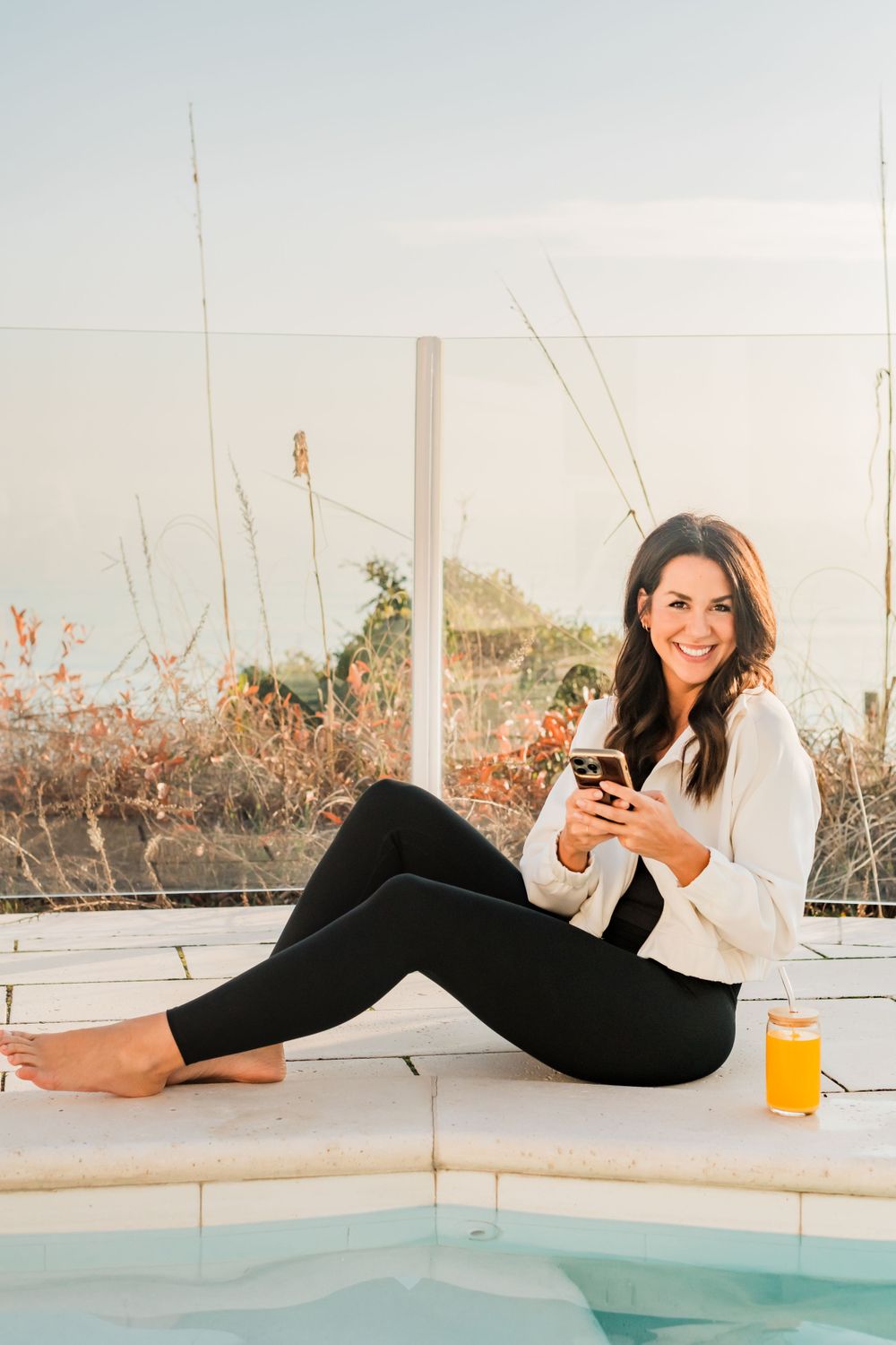 Woman relaxing by pool with phone and orange juice on sunny afternoon.