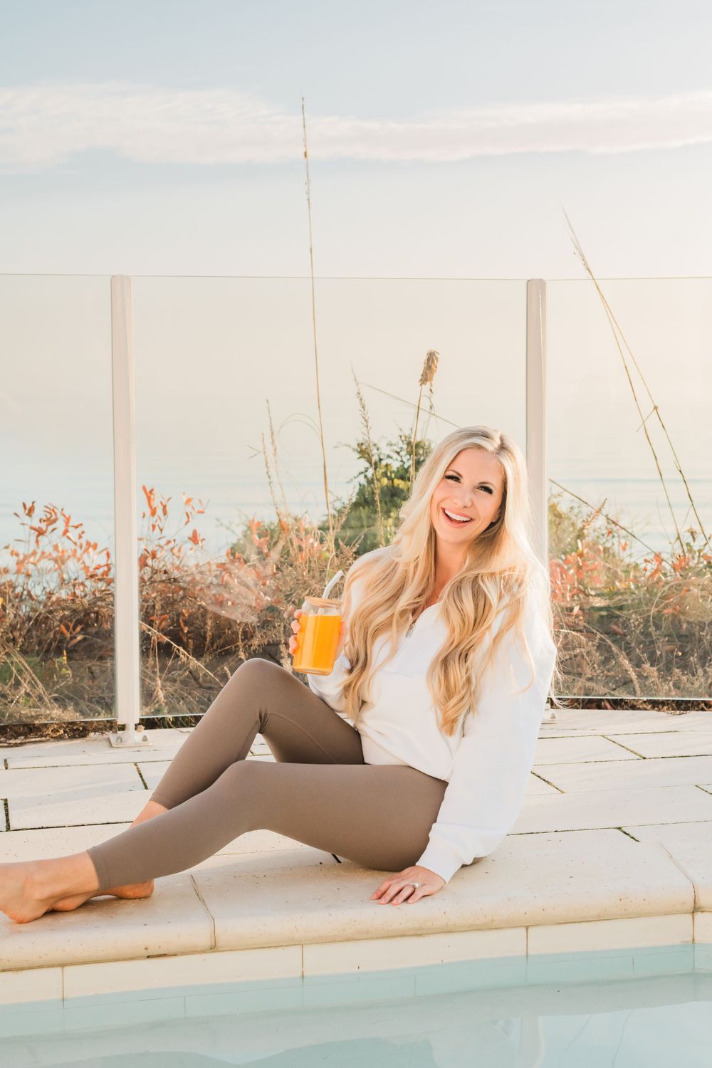 Woman in white sweater and leggings relaxes poolside with orange drink during sunset.