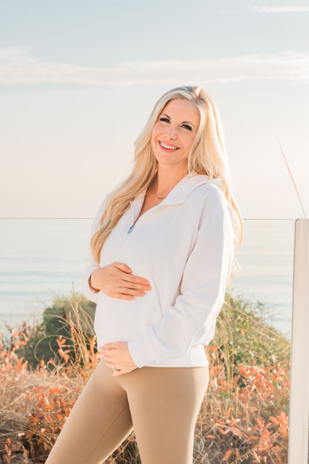 Expectant figure in white sweater poses by coastline with ocean view and coral-colored wildflowers at sunset.