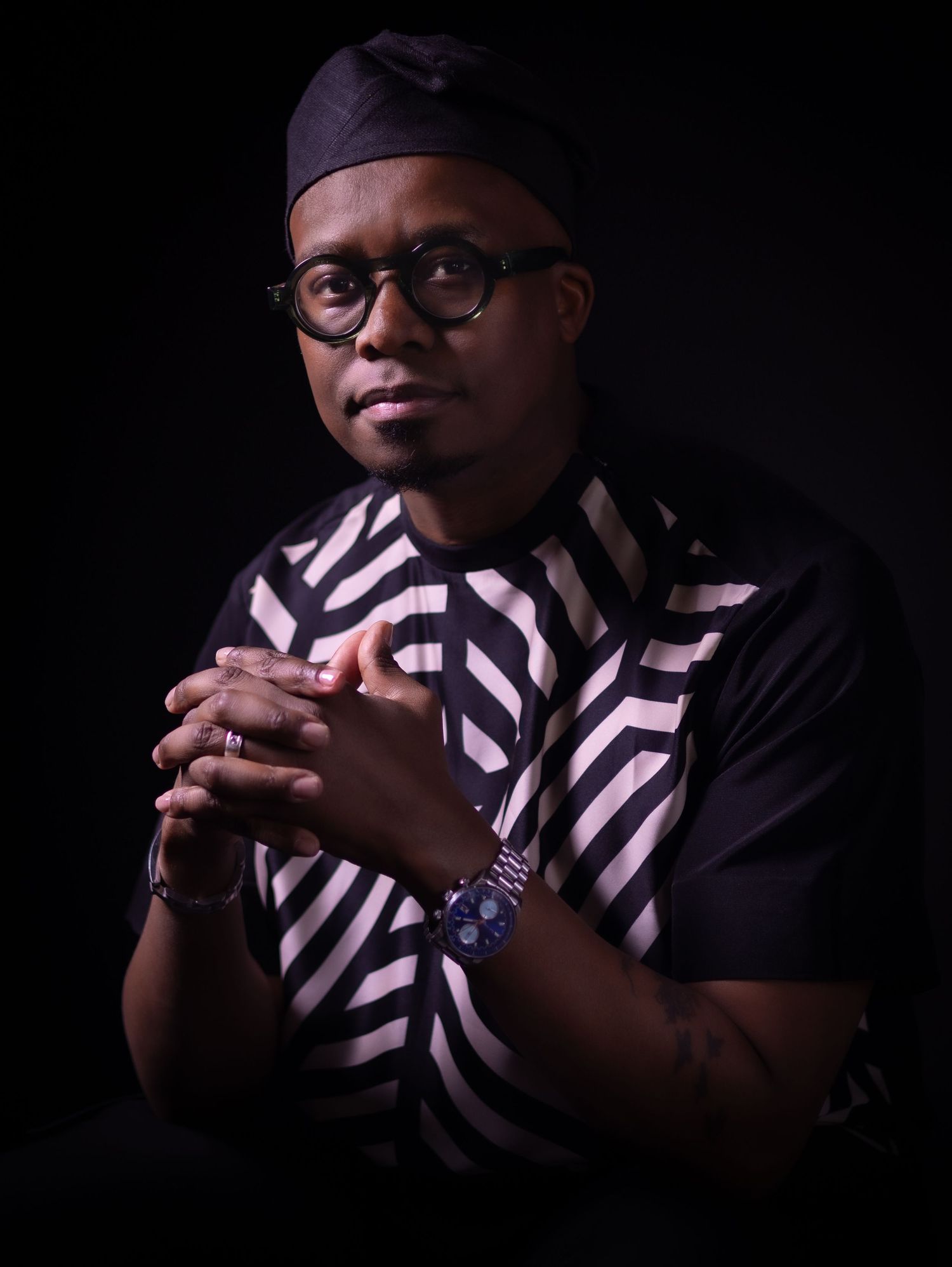 Portrait in dramatic lighting featuring a person wearing glasses, a black cap and striped shirt against dark background.