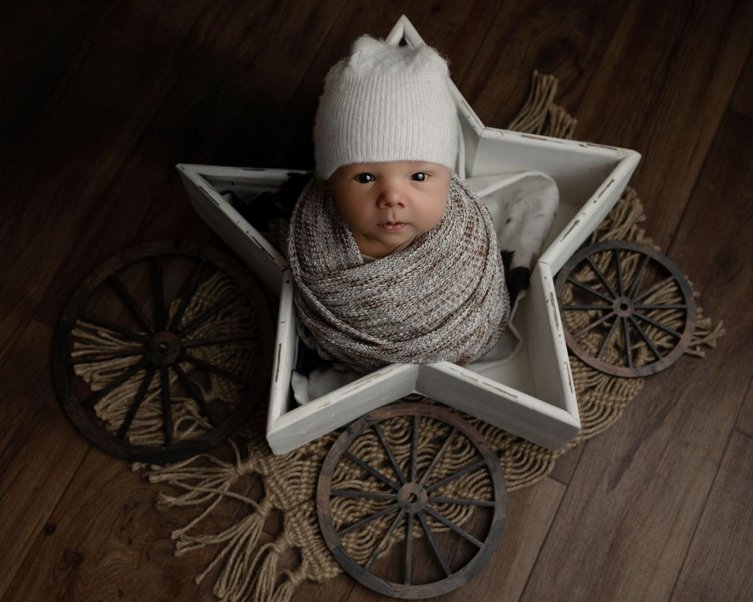 Baby wrapped in gray knit blanket sits in white star-shaped prop on wooden floor with decorative background.