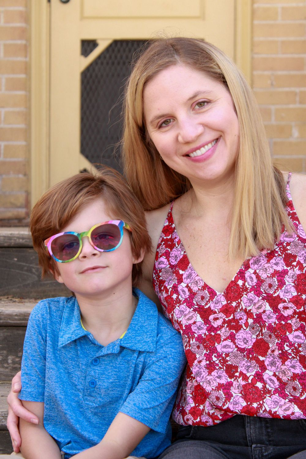 A child wearing colorful sunglasses and a blue shirt sits on steps with someone in a red floral top at a brick building.